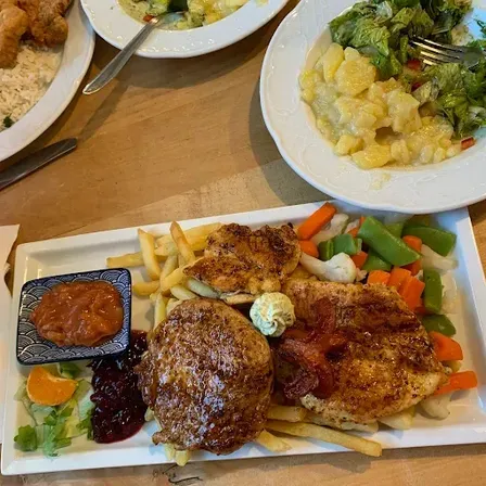 Ein Holztisch hat ein Hauptgericht mit Hühnchen, Speck, Gemüse und Saucen. Zwei weitere Teller enthalten Salat und Reis.