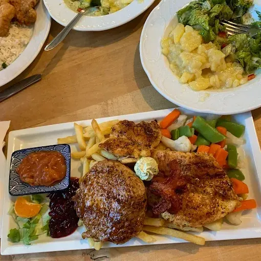 Ein Holztisch hat ein Hauptgericht mit Hühnchen, Speck, Gemüse und Saucen. Zwei weitere Teller enthalten Salat und Reis.