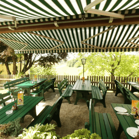 Ein Restaurant im Freien mit grünen Tischen und Bänken unter einem gestreiften Vordach, umgeben von Bäumen und einem Holzzaun.