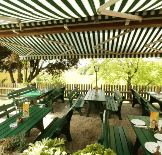 Ein Restaurant im Freien mit grünen Tischen und Bänken unter einem gestreiften Vordach, umgeben von Bäumen und einem Holzzaun.