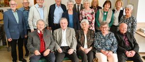 Eine Gruppe älterer Menschen, sowohl Männer als auch Frauen, sind für ein Foto versammelt. Sie sitzen auf grünen Stühlen, und einige stehen. Sie tragen alle formelle Kleidung. Der Raum hat Holztüren und der Boden ist aus Holz.