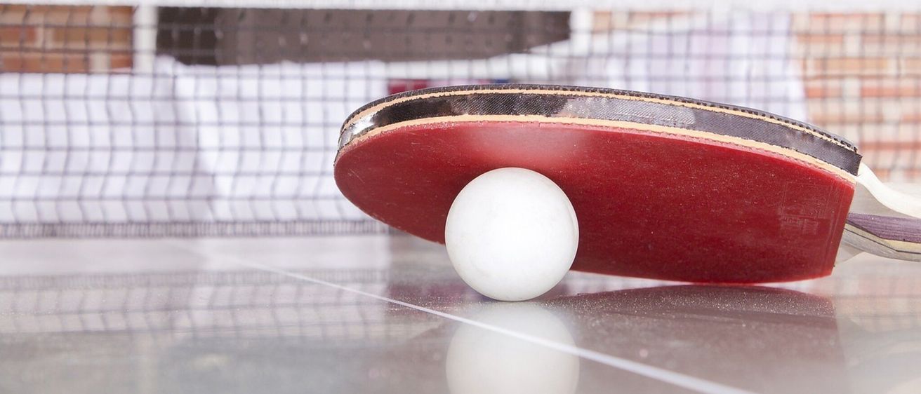 Eine Nahaufnahme eines roten Tischtennisschlägers mit einem weißen Ball auf einer reflektierenden Oberfläche. Im Hintergrund ein verschwommenes Netz und eine Ziegelwand.