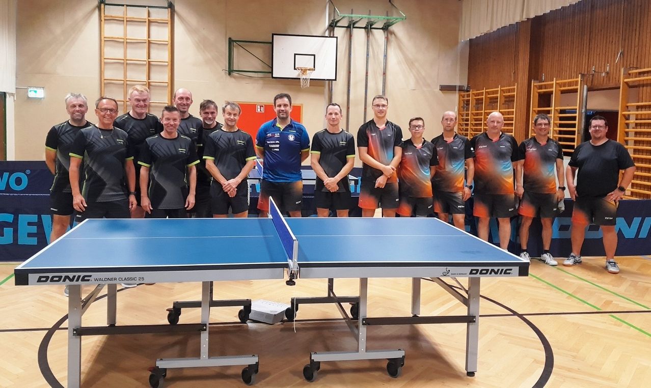 Eine Gruppe von Männern in Sportbekleidung steht hinter zwei Tischtennistischen in einer Turnhalle. Ein Mann in einem blauen Shirt lächelt.