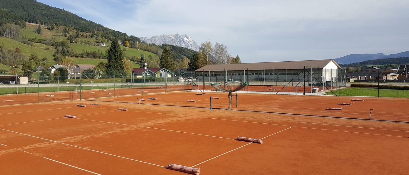 Ein leerer Tennisplatz mit rotem Sand und weißen Markierungen. Berge und Häuser im Hintergrund.