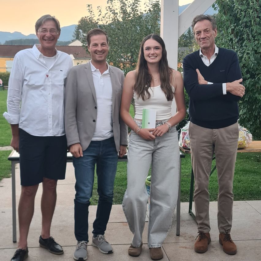 Vier Personen stehen zusammen draußen. Eine Frau hält ein Buch. Hinter ihnen steht ein Tisch mit Tüten. Bäume und Berge sind im Hintergrund.