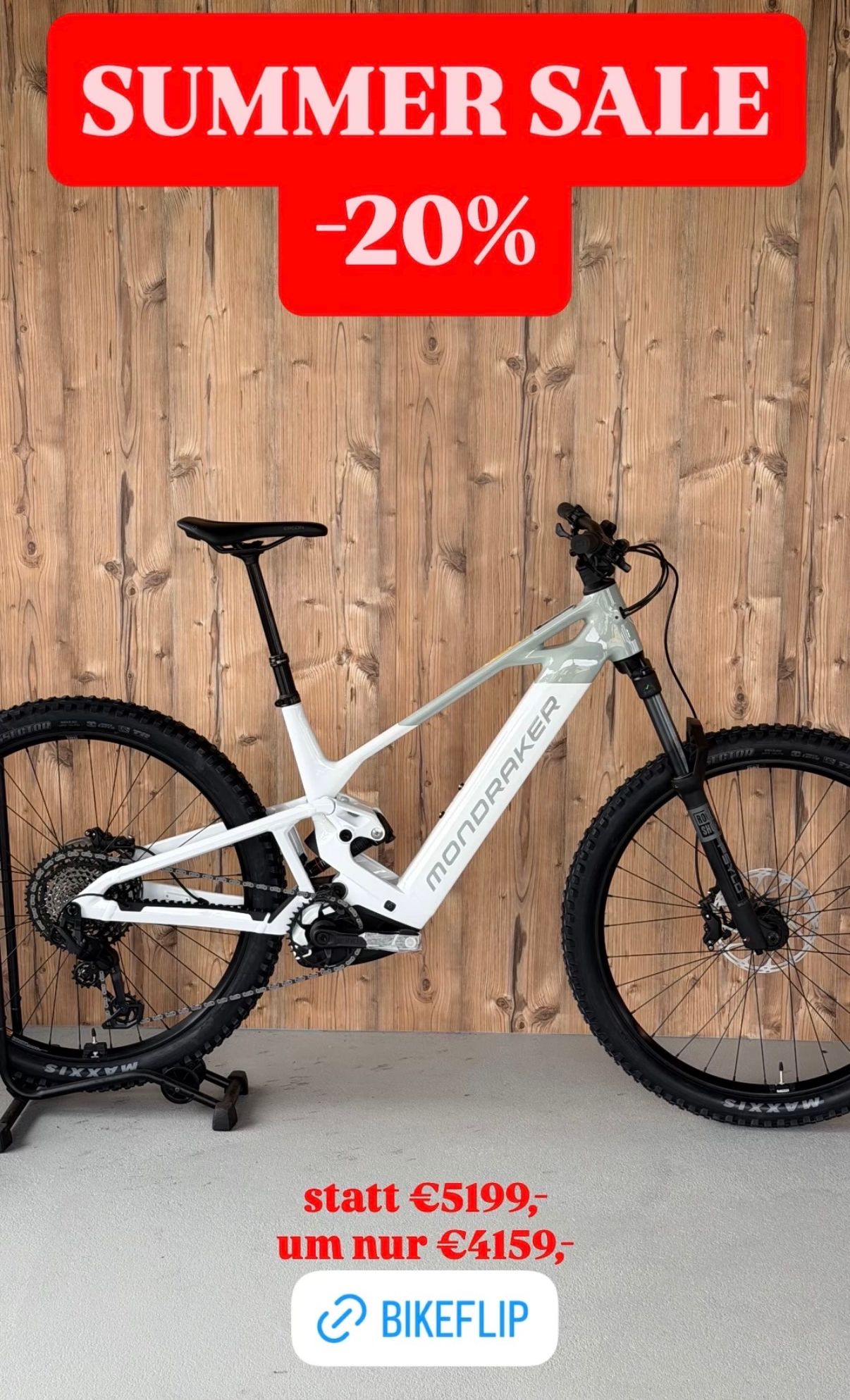 A white Mondraker mountain bike is displayed against a wooden wall, showcasing its sleek design and robust tires.