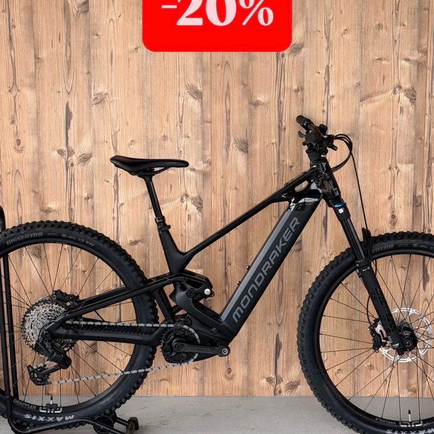 A black Mondraker mountain bike is displayed against a wooden wall with a 20% discount sign. The bike features a sleek design, a black seat, and a silver chain.