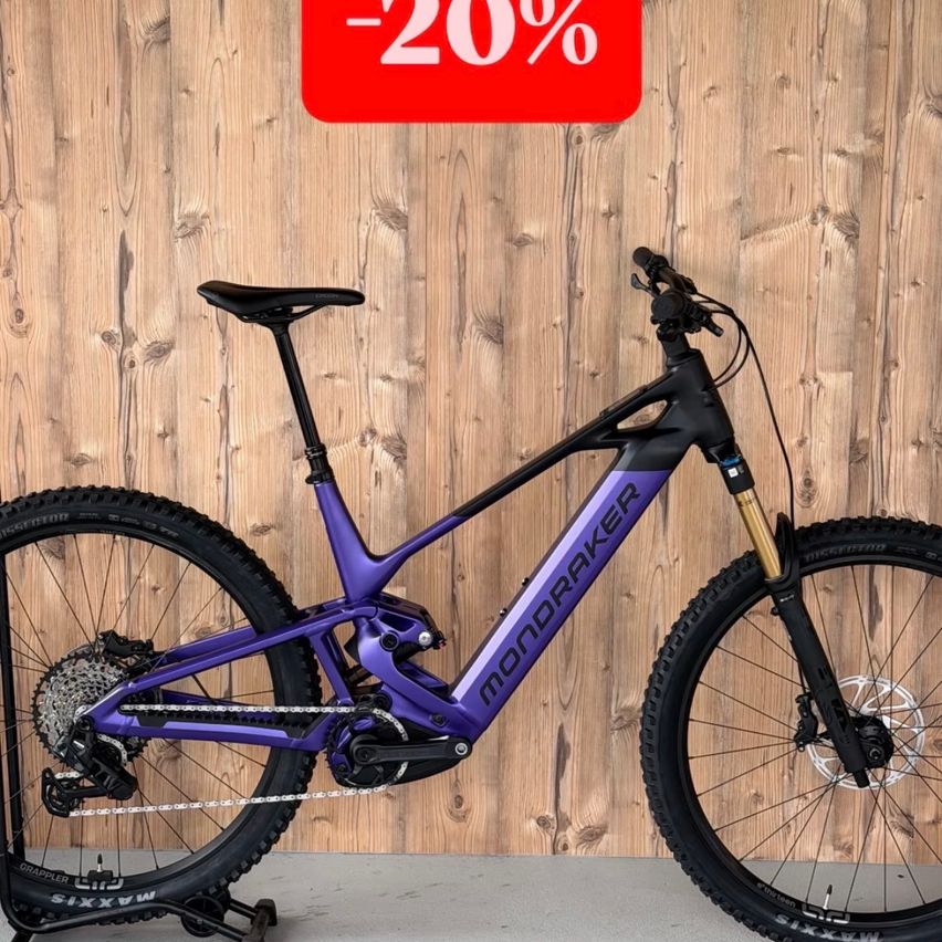 A purple and black Mondraker mountain bike is displayed against a wooden wall with a red sign indicating a 20% discount. The bike has a sleek design, with a black seat and handlebars.