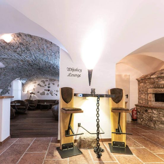 Das Innere einer Whiskey-Lounge zeigt zwei hohe Hocker mit Ketten, die an einem zentralen Tisch befestigt sind. Der Raum hat eine Steindecke und Wände mit einem rustikalen Kamin rechts. Eine Lampe beleuchtet den Bereich, und ein Schild an der Wand lautet 'Whiskey Lounge'.