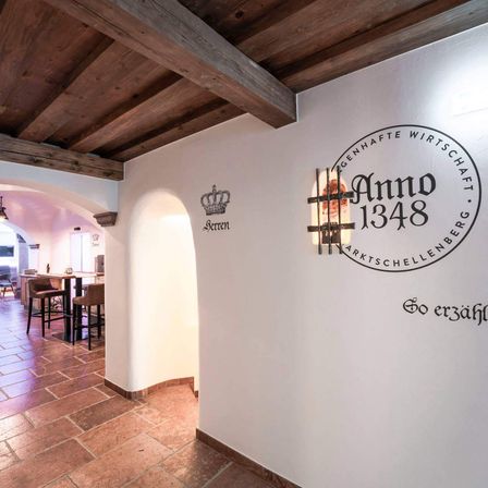 Ein Innenansicht eines Restaurants mit einer Holzdecke, einer weißen Wand und einem runden Schild mit der Aufschrift 'Anno 1348'. Es gibt einen Tisch und Stühle, eine Lampe und ein Fenster auf der linken Seite.