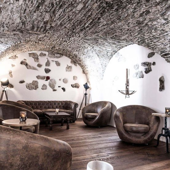 Ein eleganter Loungebereich mit Holzböden und Steinwänden, bestehend aus einer Couch und runden Stühlen mit Tisch, ergänzt durch Lampen und ein dekoratives Metallstück an der Wand.