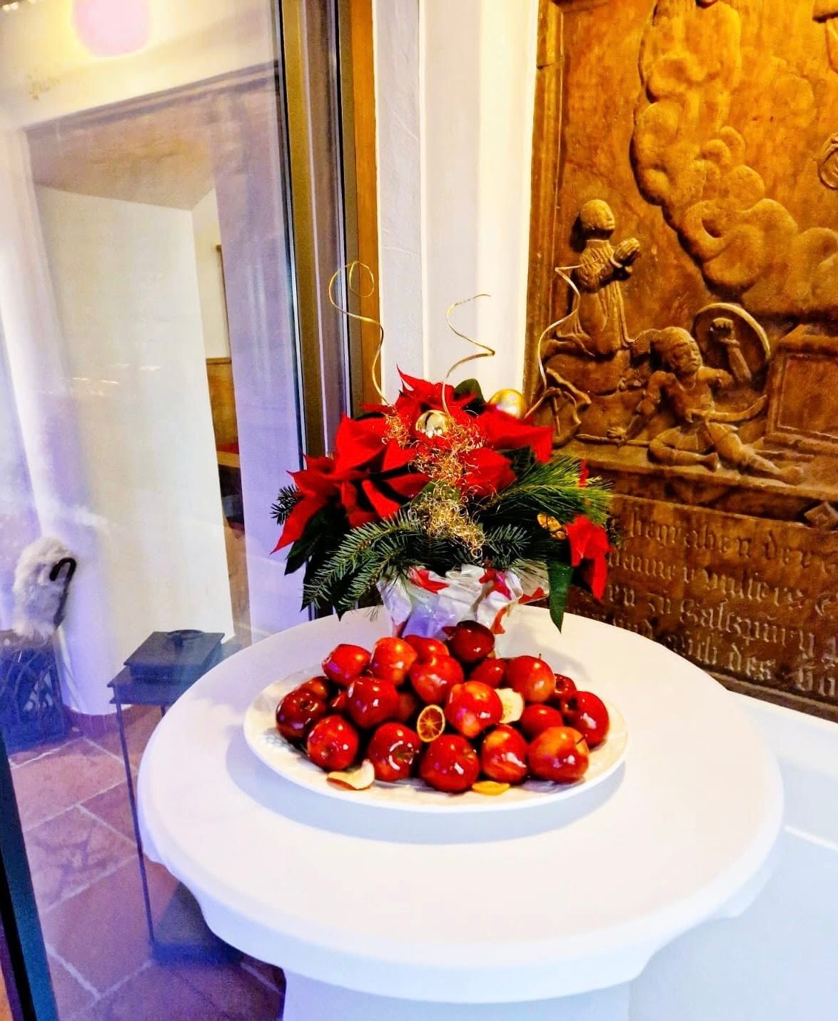 Ein weißer Tisch zeigt einen Teller mit roten Äpfeln, begleitet von einer Blumenarrangement aus roten Blumen und Grünpflanzen. Der Hintergrund zeigt ein holzgeschnitztes Kunstwerk mit Figuren und Text.