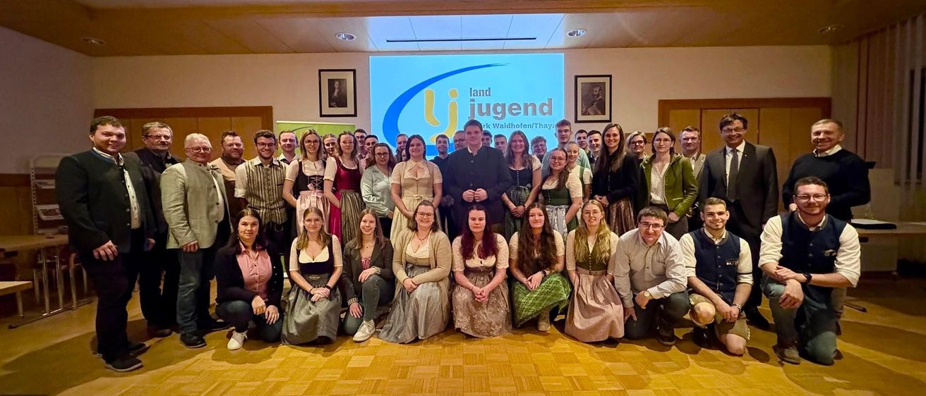 Eine Gruppe von Menschen in traditioneller Kleidung, die für ein Foto in einem Saal posiert. Hinter ihnen befindet sich eine Projektionsleinwand mit dem 'Land Jugend' Logo.