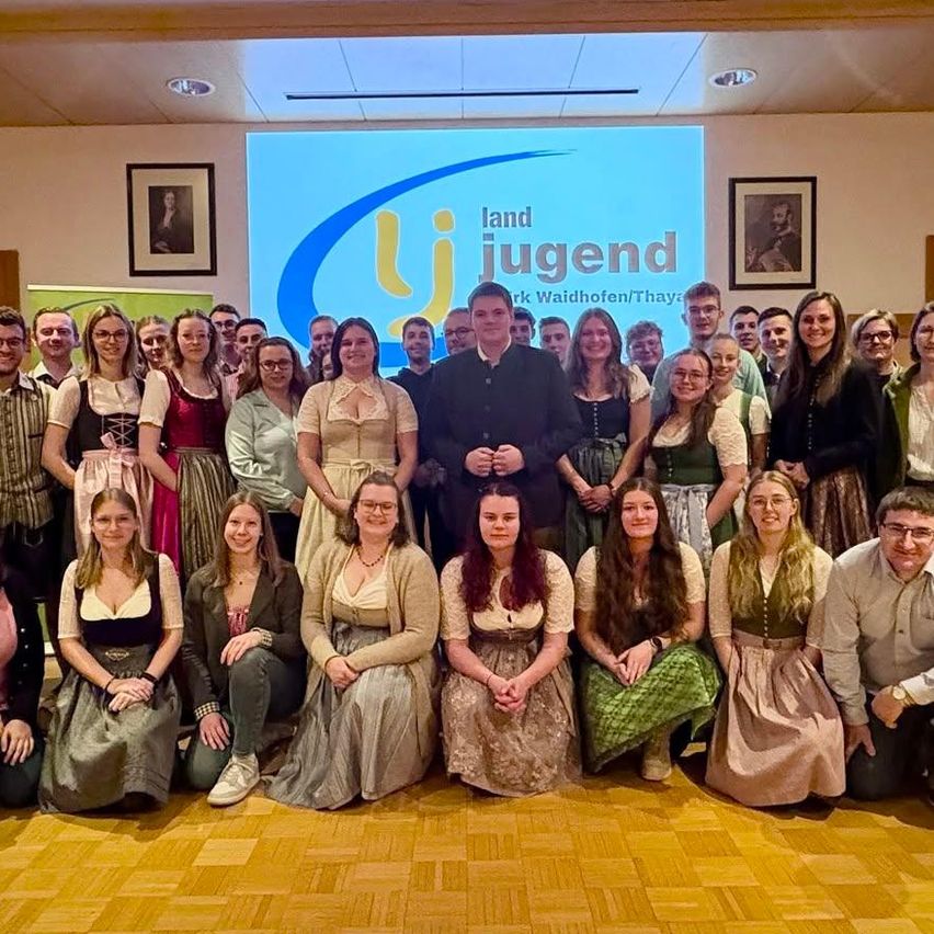 Eine Gruppe von Menschen in traditioneller Kleidung, die für ein Foto in einem Saal posiert. Hinter ihnen befindet sich eine Projektionsleinwand mit dem 'Land Jugend' Logo.