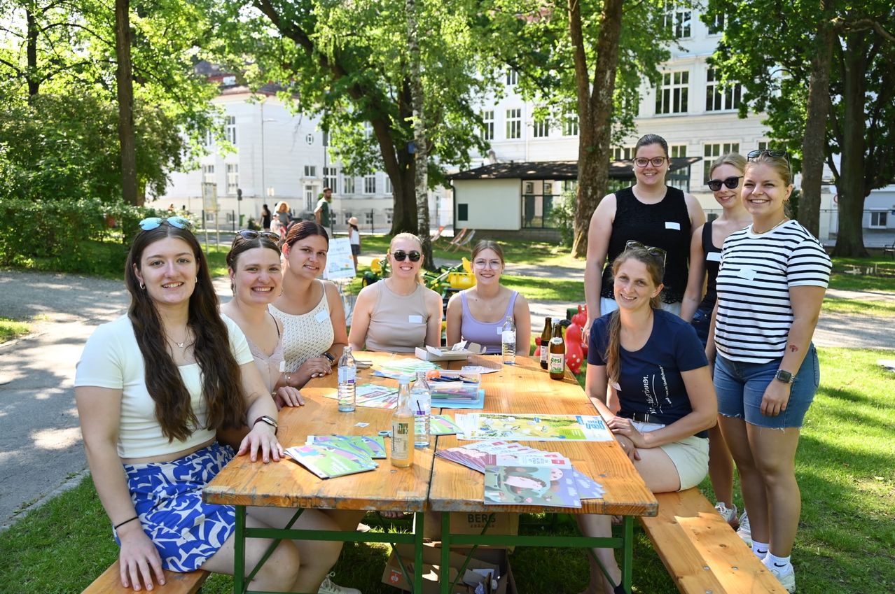 Eine Gruppe von Frauen sitzt um einen Tisch in einem Park. Sie lächeln und posieren für ein Foto. Auf dem Tisch befinden sich Flaschen, Papiere und ein Korb. Die Frauen tragen Freizeitkleidung und einige tragen Sonnenbrillen. Hinter ihnen sind Bäume und Gebäude.