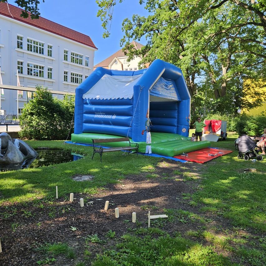 Ein blauer und grüner aufblasbarer Hüpfburg steht in einem Park, mit einer Bank und einem roten Teppich davor. Ein kleiner Teich und ein Gebäude mit vielen Fenstern dahinter.