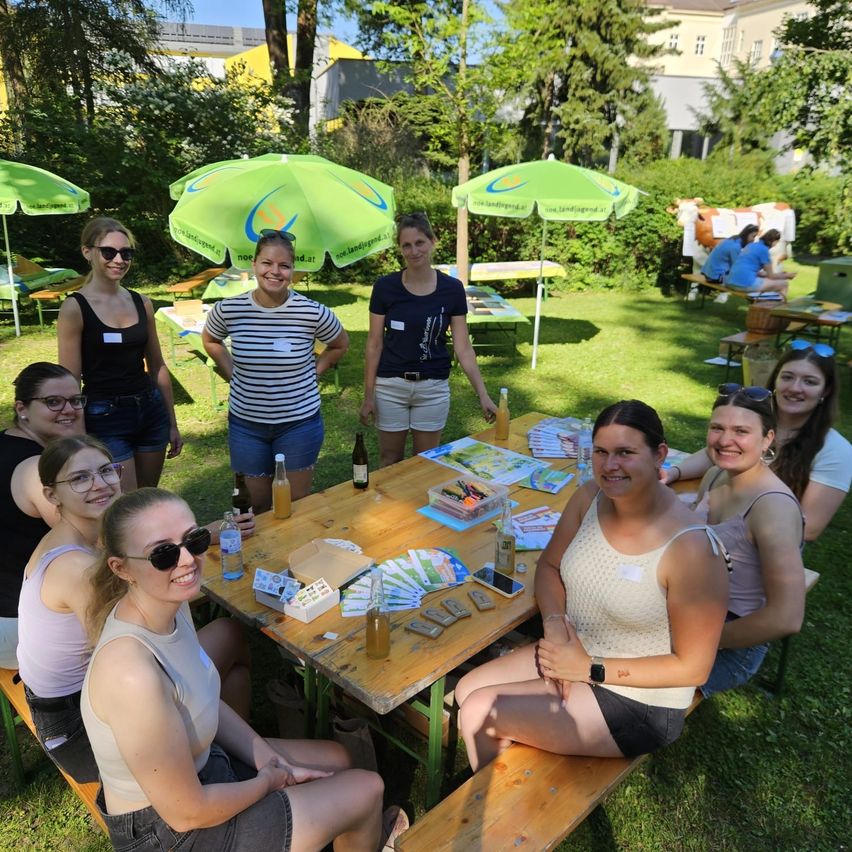 Eine Gruppe von Frauen sitzt um einen Tisch in einem Park und genießt Getränke und Snacks. Sie sind lässig gekleidet, einige tragen Sonnenbrillen und Armbänder. Der Tisch ist mit Tellern, Getränken und anderen Gegenständen gedeckt. Im Hintergrund befinden sich grüne Sonnenschirme und ein Gebäude.