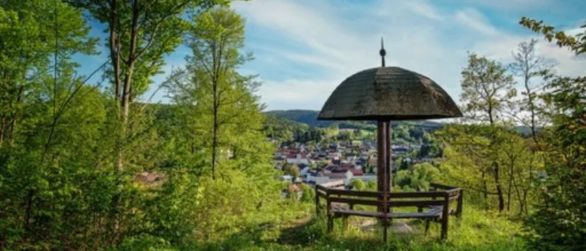 Ein Pavillon auf einem grasbewachsenen Hügel bietet einen Blick auf ein Dorf, eingebettet im Tal, umgeben von Bäumen unter einem teilweise bewölkten Himmel.