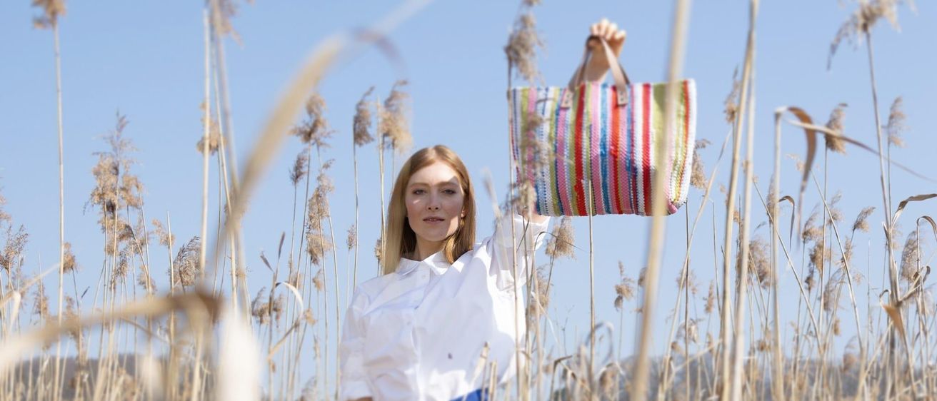 Eine Frau steht in einem hohen Feld und hält eine bunt gestreifte Tasche. Sie trägt ein weißes Hemd und blaue Hosen. Der Himmel ist klar und blau.