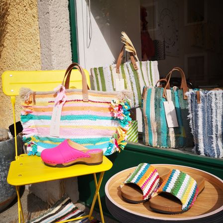 Ein gelber Stuhl zeigt einen rosa Schuh und eine farbenfrohe gewebte Tasche. Dahinter sind drei Handtaschen auf einem grünen Regal ausgestellt. Die Taschen sind mehrfarbig und gewebt.