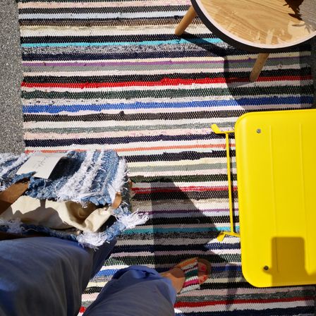 Ein bunter gestreifter Teppich bedeckt den Boden mit einem hölzernen Hocker und einem gelben Koffer darauf. Eine Person in Hausschuhen steht auf dem Teppich.