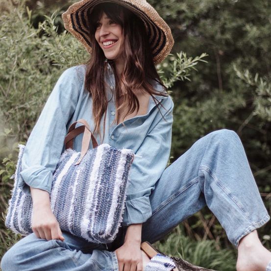 Eine Frau in Hut und Jeans sitzt auf einem Baumstumpf und lächelt, mit einer gehäkelten Tasche im Schoß.