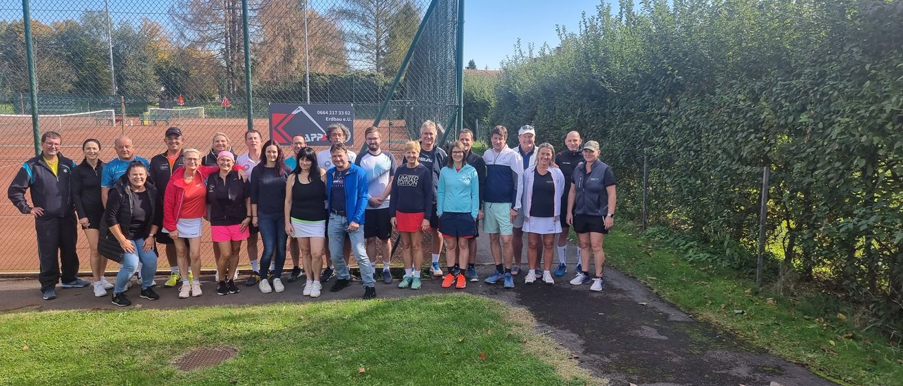Eine Gruppe von Menschen posiert für ein Foto vor einem Tennisplatz. Der Platz ist von einem Zaun umgeben. Die Menschen tragen Turnschuhe und einige von ihnen tragen Hüte.