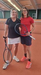 Zwei Frauen posieren mit Tennisschlägern auf einem Sandplatz, eine in Schwarz und eine in Rot.