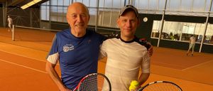Zwei Männer stehen auf einem Tennisplatz, beide halten Tennisschläger. Der Mann auf der linken Seite trägt ein blaues Shirt und graue Shorts. Der Mann auf der rechten Seite trägt ein weißes Shirt und schwarze Shorts. Beide lächeln.