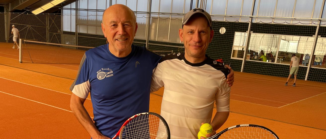 Zwei Männer stehen auf einem Tennisplatz, beide halten Tennisschläger. Der Mann auf der linken Seite trägt ein blaues Shirt und graue Shorts. Der Mann auf der rechten Seite trägt ein weißes Shirt und schwarze Shorts. Beide lächeln.