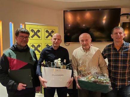 Vier Männer stehen nebeneinander in einem Raum. Zwei von ihnen halten Geschenkboxen, eine mit Weinflaschen und die andere mit verschiedenen Gegenständen. Sie lächeln und scheinen ein Ereignis zu feiern. Hinter ihnen steht ein großer Monitor und ein gelber Banner mit schwarzen Kreuzen.