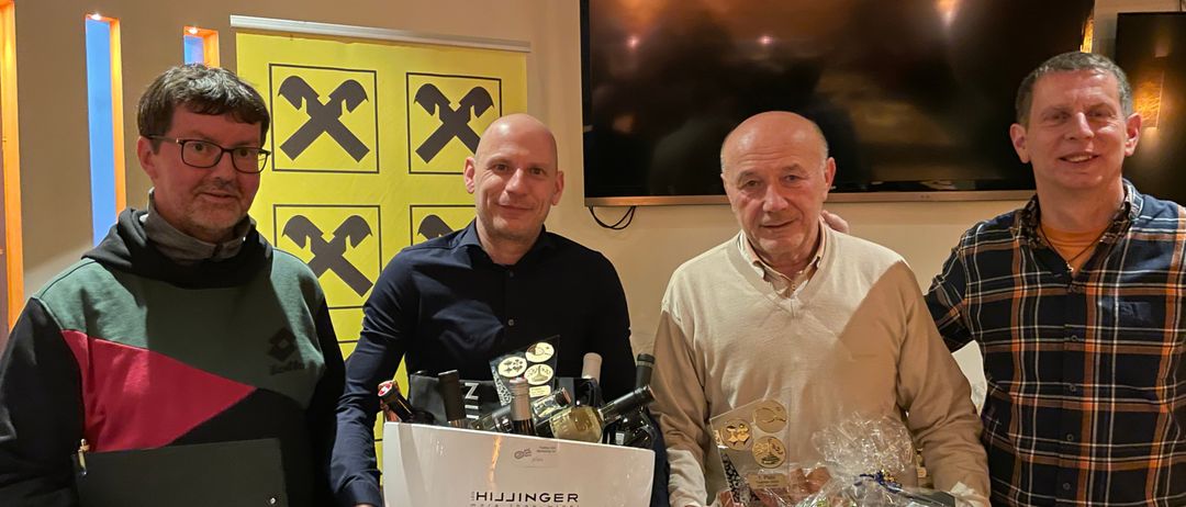 Vier Männer stehen lächelnd in einem Raum zusammen. Der Mann auf der linken Seite hält eine Mappe. Der Mann in der Mitte hält eine weiße Box mit Flaschen darin. Der Mann auf der rechten Seite hält eine grüne Box mit einem Geschenk darin. Dahinter befindet sich ein gelber Banner mit dem Buchstaben X.