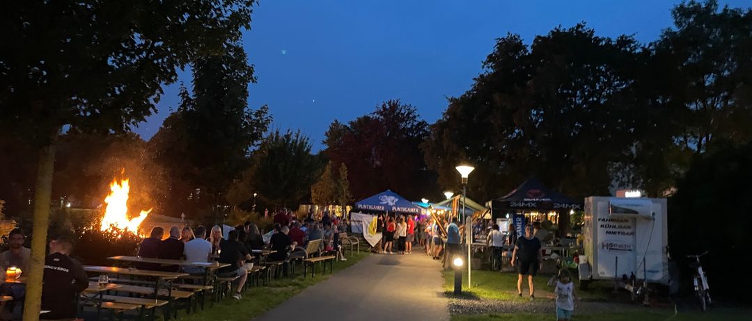 Bei Dämmerung versammeln sich Menschen in einem Park, einige sitzen an Picknicktischen in der Nähe eines Lagerfeuers. Straßenlaternen und Zelte säumen den Weg. Ein Kind rennt im Vordergrund.