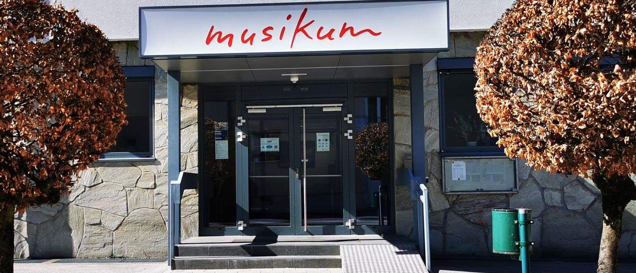 Ein Gebäude mit Glastüren und Fenstern, mit einem Namensschild, das 'musikum' liest. Es gibt Pflanzen auf beiden Seiten des Eingangs.