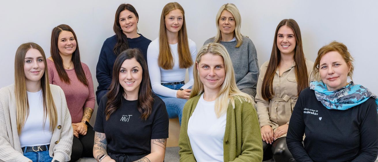 Fünf Frauen sitzen auf einer Bank und lächeln alle. Eine Frau vorne trägt ein schwarzes Shirt mit weißem Text. Eine andere Frau trägt einen grünen Cardigan.