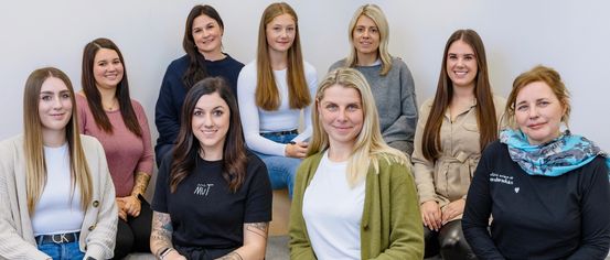 Fünf Frauen sitzen auf einer Bank und lächeln alle. Eine Frau vorne trägt ein schwarzes Shirt mit weißem Text. Eine andere Frau trägt einen grünen Cardigan.