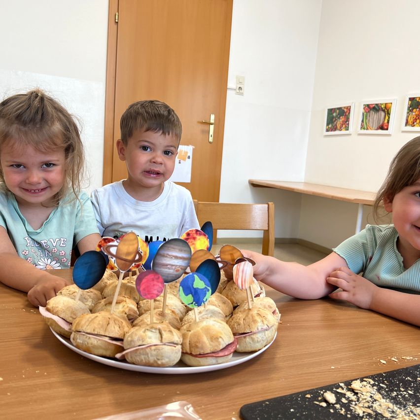 Drei Kinder sitzen an einem Tisch mit einem Teller Sandwiches, jeweils mit einem Planetenaufkleber. Sie scheinen zu lächeln und genießen ihr Essen. Hinter ihnen befindet sich eine Holztür und einige gerahmte Bilder an der Wand.