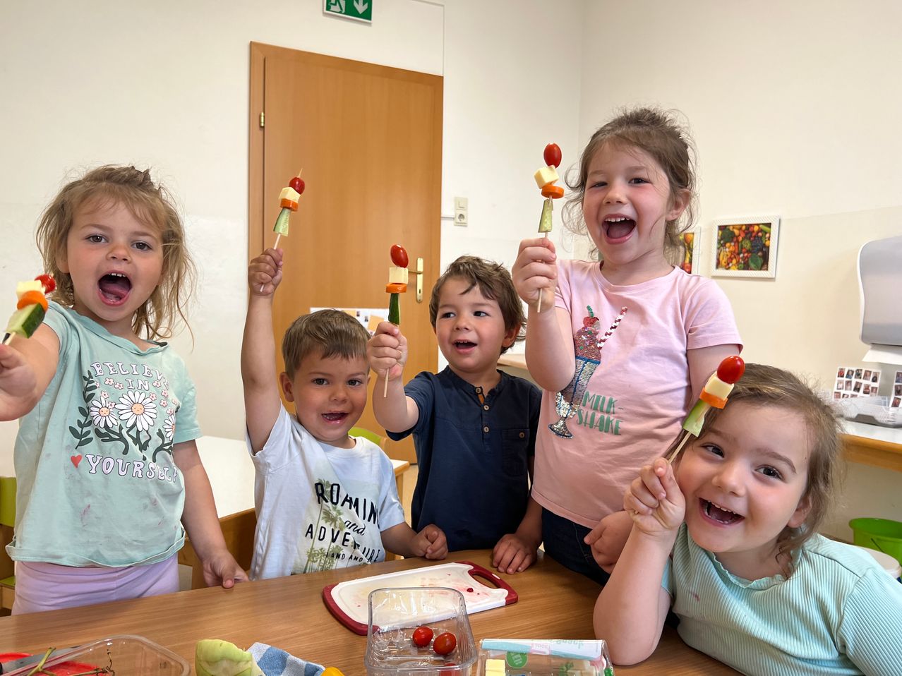Fünf Kinder in einem Klassenzimmer, die Obstspieße hochhalten. Sie lächeln und posieren für ein Foto.