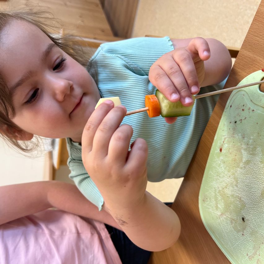 Ein kleines Mädchen macht einen Obstspieß mit einer Gurkenscheibe und einer Karottenstange.
