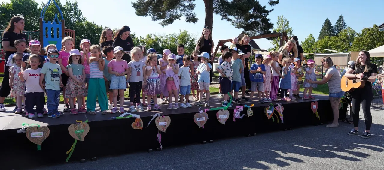 Eine Gruppe von Kindern steht auf einer Bühne mit Herzdekorationen, einige tragen Hüte, und Erwachsene stehen hinter ihnen. Bäume und eine Schaukel sind im Hintergrund.