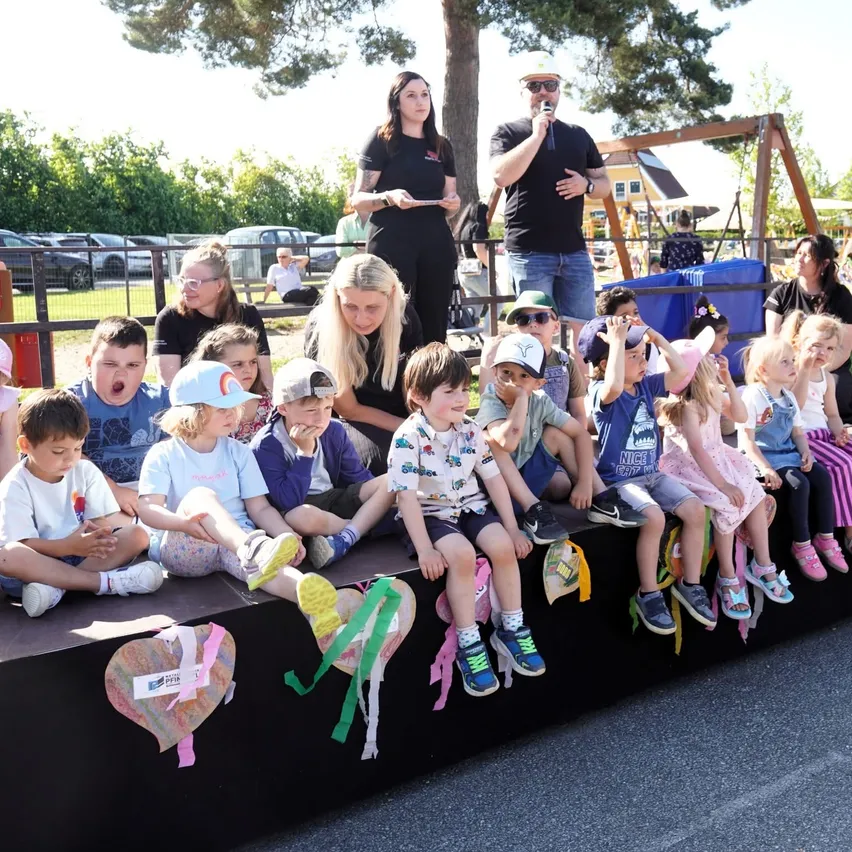 Eine Gruppe von Kindern sitzt auf einer Bühne mit herzförmigen Dekorationen. Zwei Erwachsene stehen hinter ihnen, einer hält ein Mikrofon. Eine Frau in der Front gestikuliert, und mehrere Menschen sitzen und stehen herum.