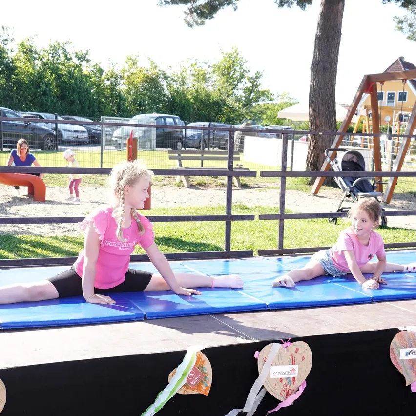 Zwei Mädchen machen Gymnastik auf einer blauen Matte auf der Bühne. Einige herzförmige Papiere sind auf der Bühne. Dahinter ist ein Metallzaun. Ein Kinderwagen steht neben dem Baum. Ein paar Autos sind hinter dem Zaun geparkt.