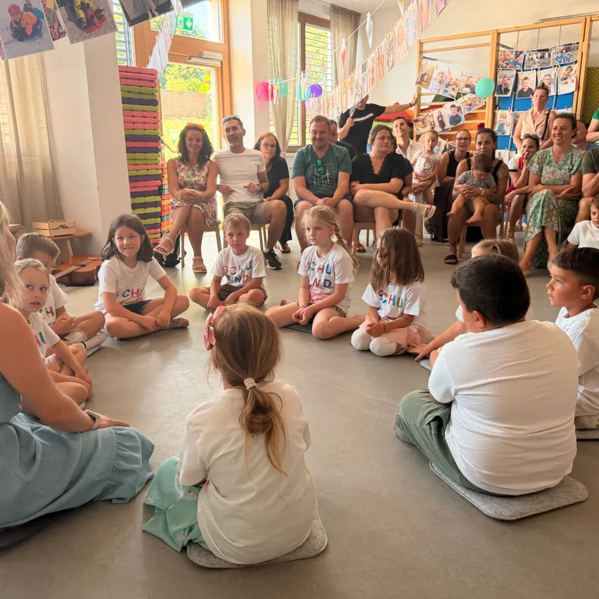 Eine Gruppe von Kindern und Erwachsenen sitzt in einem Kreis in einem Raum, wahrscheinlich einem Klassenzimmer. Die Kinder tragen weiße T-Shirts mit dem Text 'SCHULD' darauf. Die Erwachsenen sitzen auf Stühlen und einige stehen. Der Raum hat eine weiße Wand mit Bildern und einem weißen Vorhang. Es gibt auch einige Gegenstände auf dem Boden.