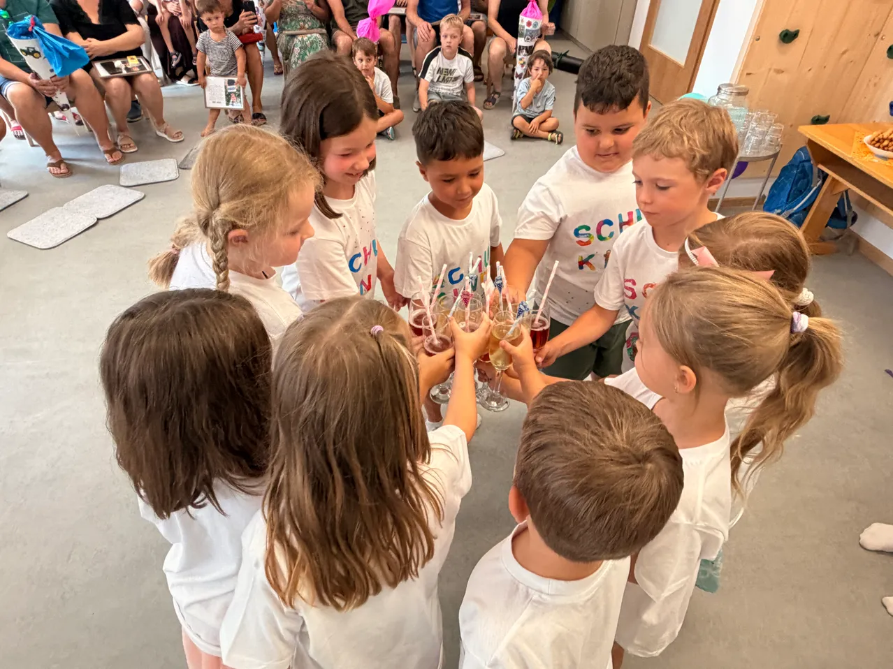 Eine Gruppe von Kindern in weißen T-Shirts mit bunten Buchstaben steht im Kreis und hält Gläser mit Saft in der Hand. Dahinter sitzen und stehen Erwachsene und Kinder in einem Raum.