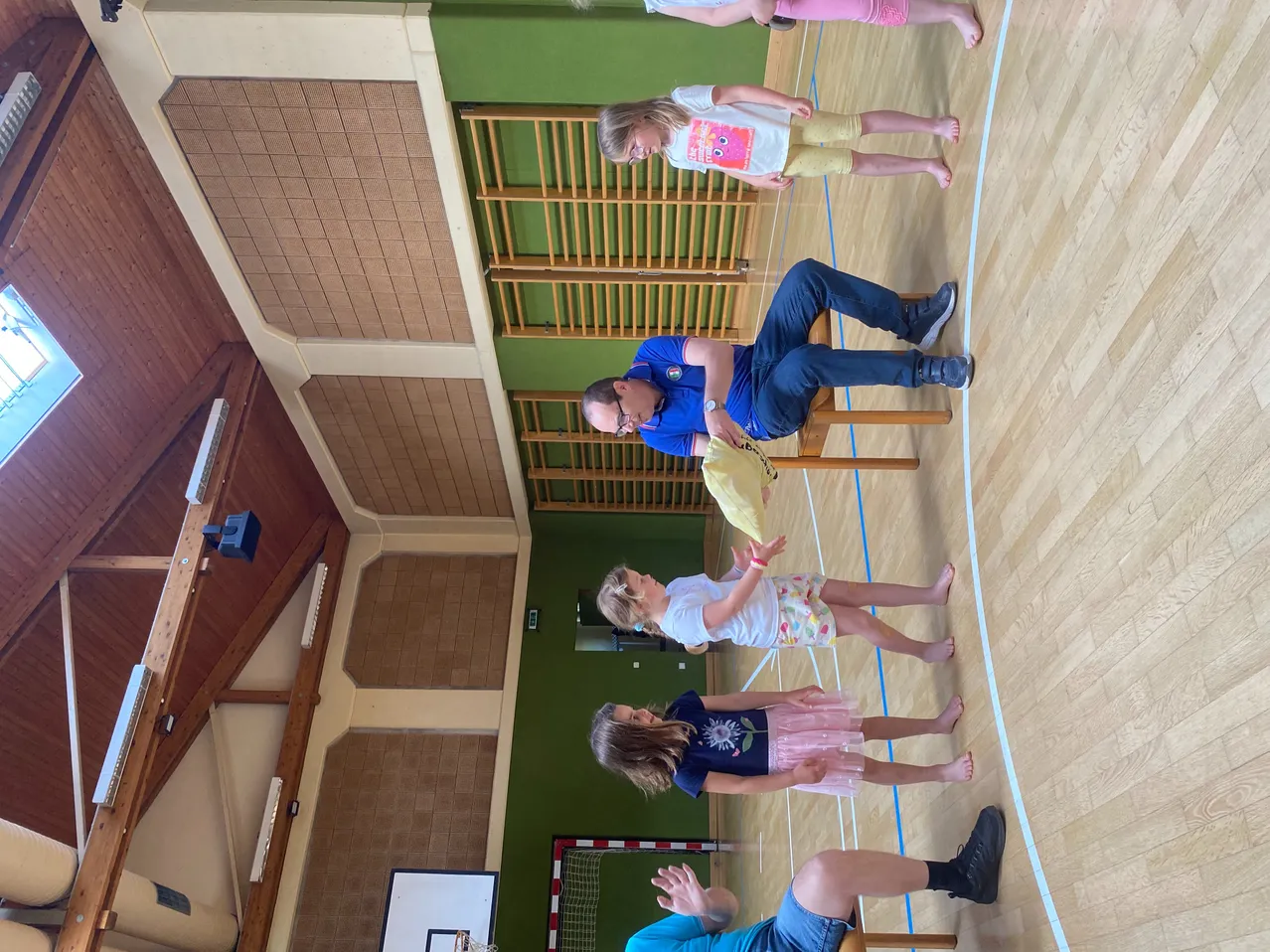 Eine Gruppe von Kindern und Erwachsenen befindet sich in einer Sporthalle. Sie spielen ein Spiel, wobei ein Erwachsener auf einem Stuhl sitzt. Die Kinder halten gelbe Objekte und stehen um den sitzenden Erwachsenen herum.