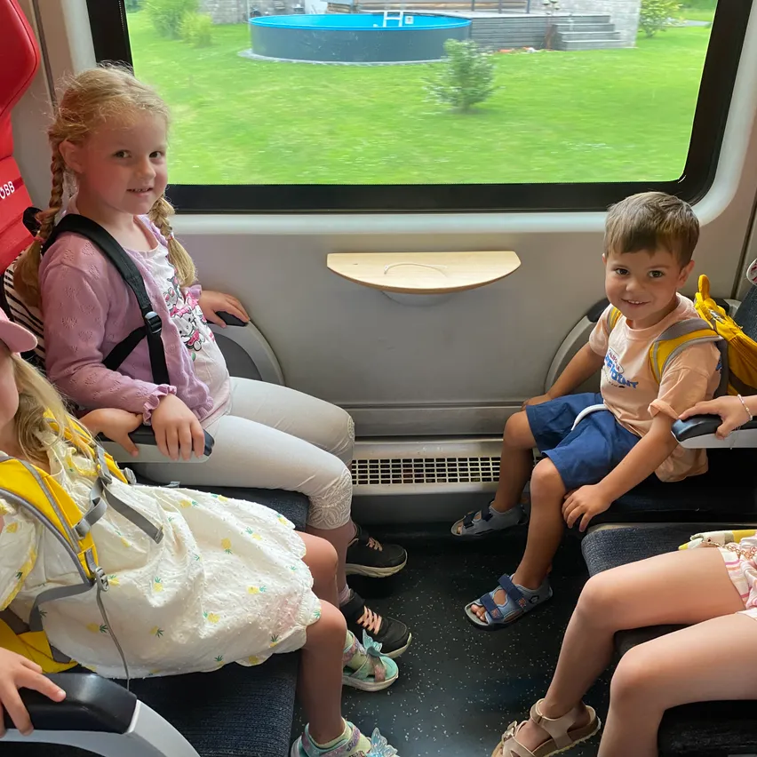 Vier Kinder sitzen in einem Zug und lächeln. Zwei Mädchen tragen Kleider, eines hat einen Rucksack. Der Junge trägt Sandalen und Shorts. Das Fenster zeigt ein grasiges Feld und einen Pool.