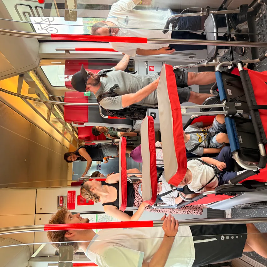 Das Innere eines überfüllten Zuges mit stehenden, sitzenden und an den Haltestangen festhaltenden Passagieren. Einige benutzen Kinderwagen.