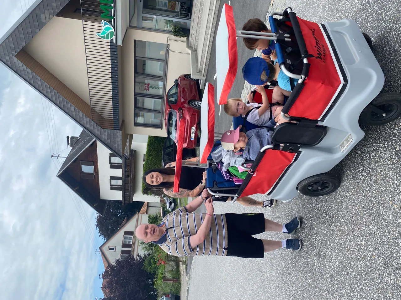 Eine Familie steht neben einem Learning Cart mit vier Kindern, zwei Erwachsenen und einem Gebäude im Hintergrund.