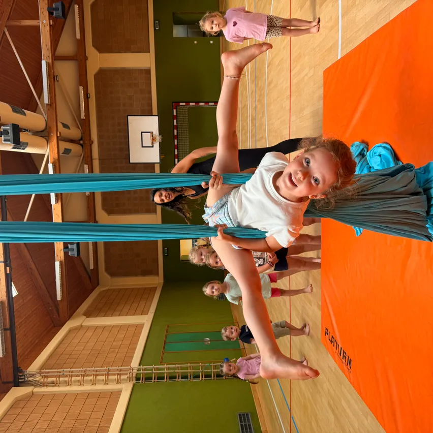 Kinder in einer Turnhalle, eines wird von blauem Stoff gehalten, lächelt. Dahinter hängt ein anderes Kind an einer Stange, ein Basketballkorb im Hintergrund.