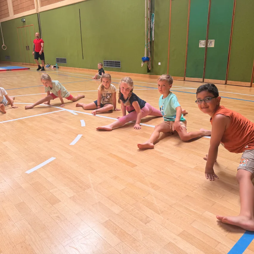 Eine Gruppe von Kindern macht Gymnastik in einer Turnhalle. Sie sitzen auf dem Boden und tragen Brillen und Turnschuhe. Ein Mann steht hinter ihnen.